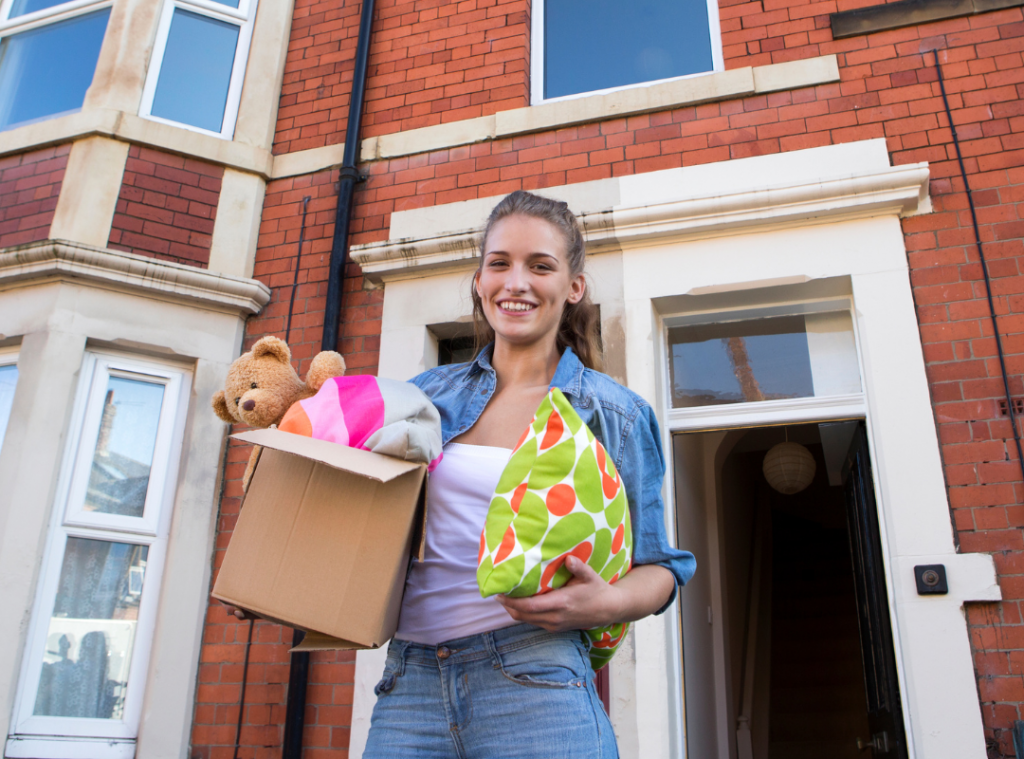 White young adult holding a box with a teddy bear and cushion moving into