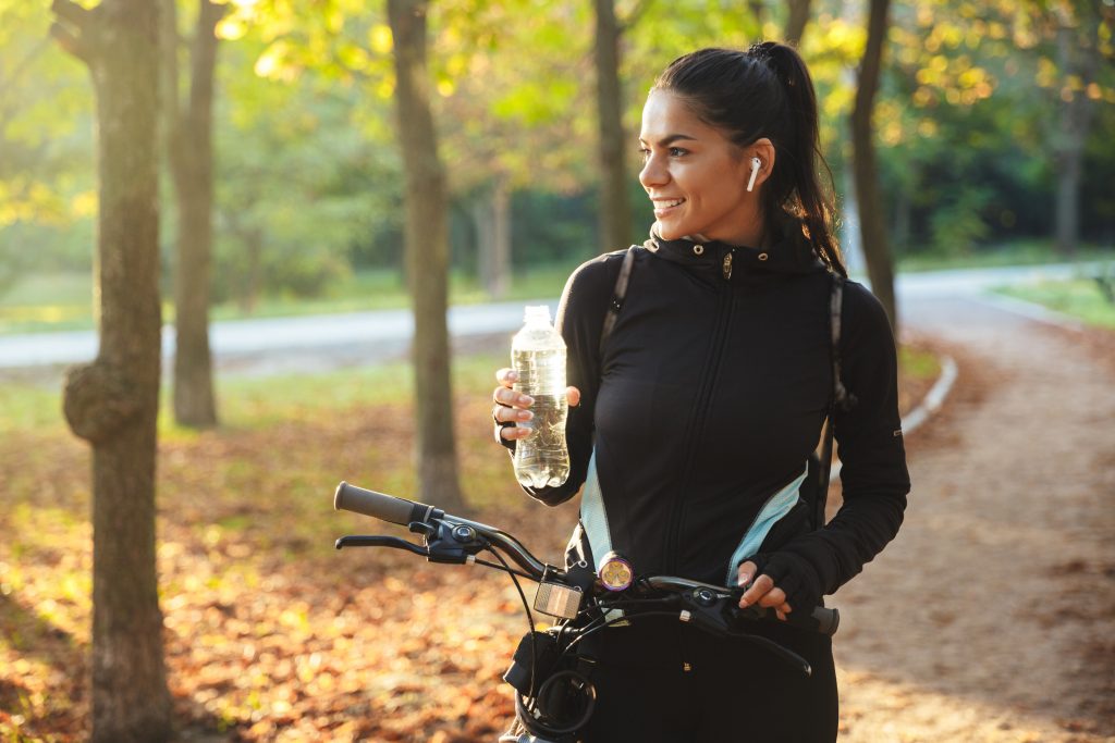 Attractive fit sportswoman with a bicycle at the park, drinking water from a bottle