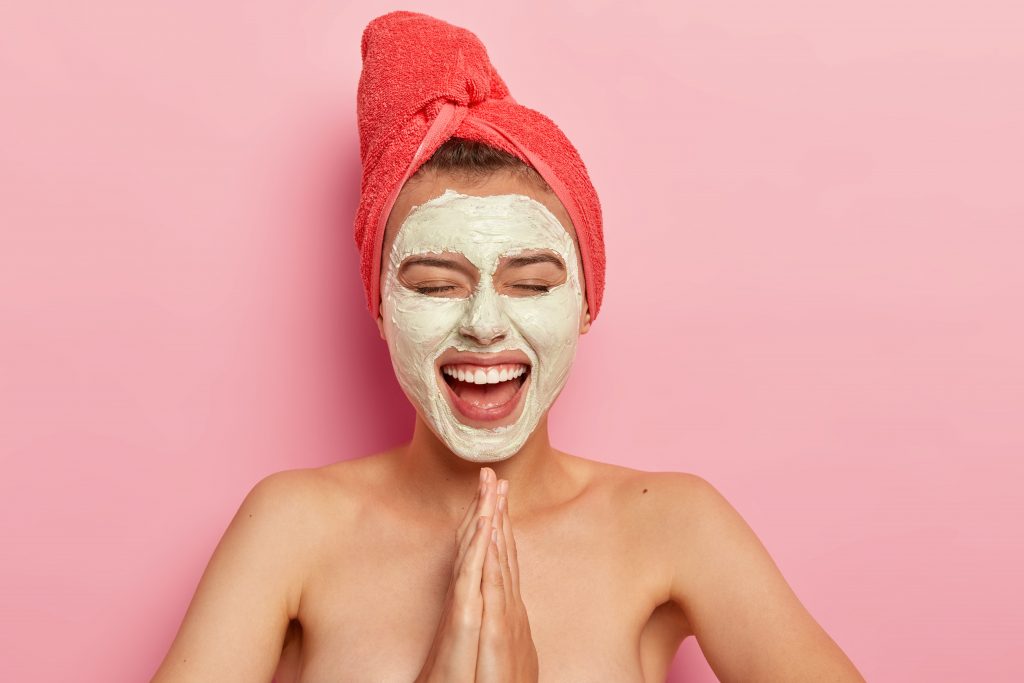 Happy pleased female keeps palms in praying gesture, smiles broadly, wears facial mask for rejuvenation, has fun at home, poses naked with bare shoulders, isolated on pink wall. Purifying, spa