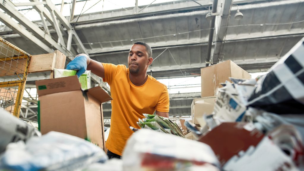 Young african american man packing boxes with waste paper for reuse while working on rubbish station, widescreen. Garbage sorting and recycling concept