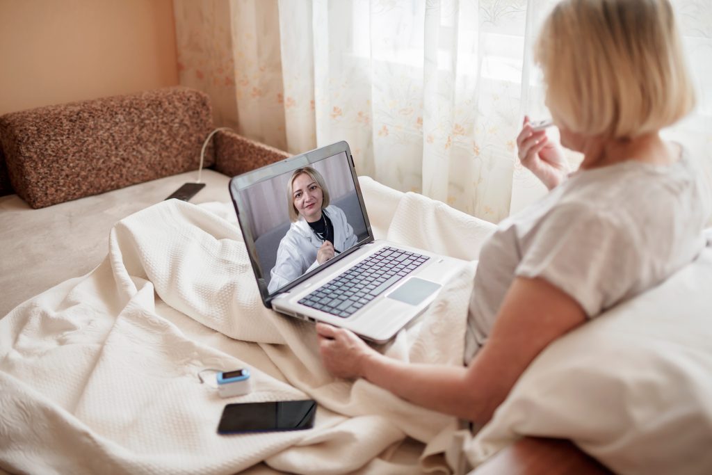 Old woman in bed looking at screen of laptop and consulting with a doctor online at home, telehealth services during lockdown, distant video call, modern tech healthcare application