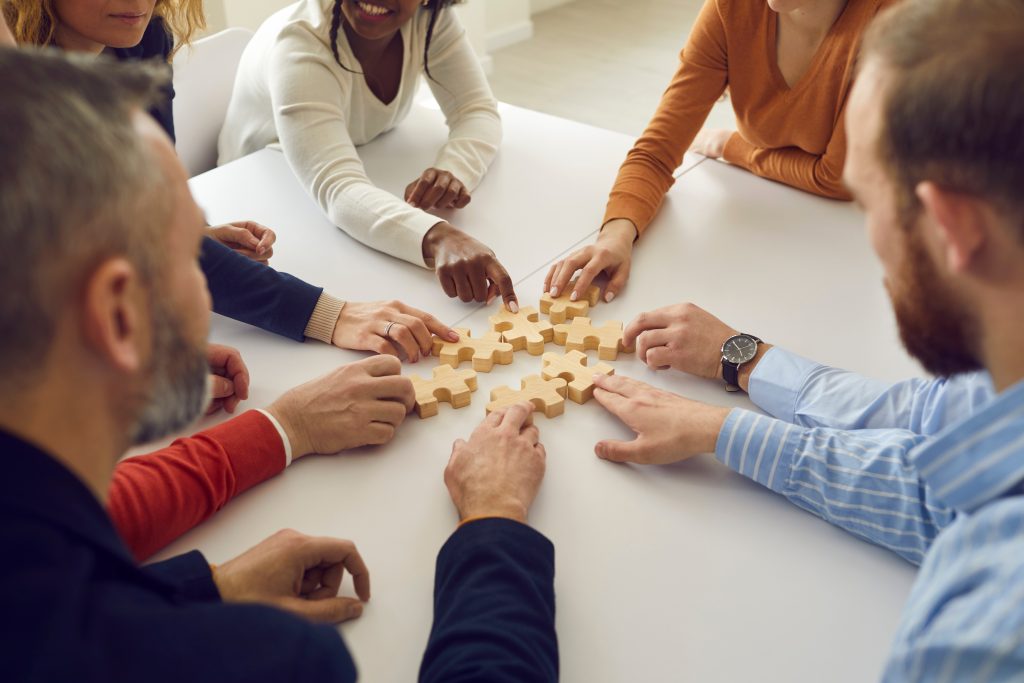 Diverse team of business people sitting around table and joining jigsaw puzzle pieces
