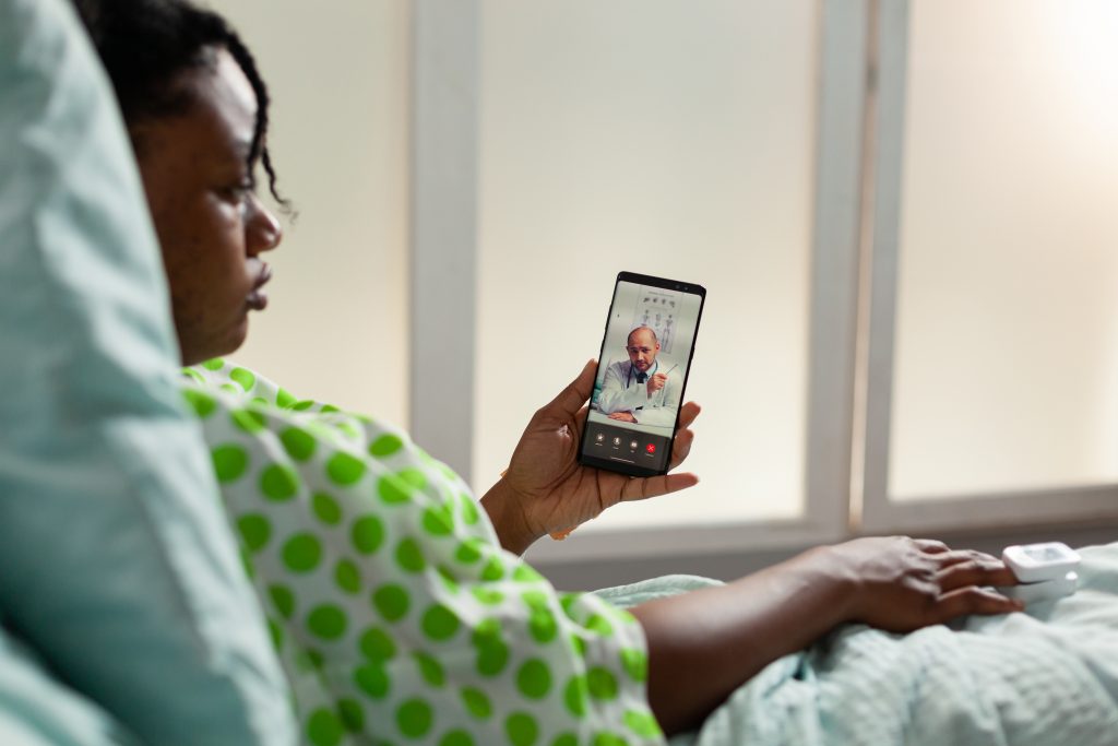 African american sick patient holding phone discussing disease expertise with remote therapist doctor during online videocall meeting conference in hospital office. Telehealth call on screen