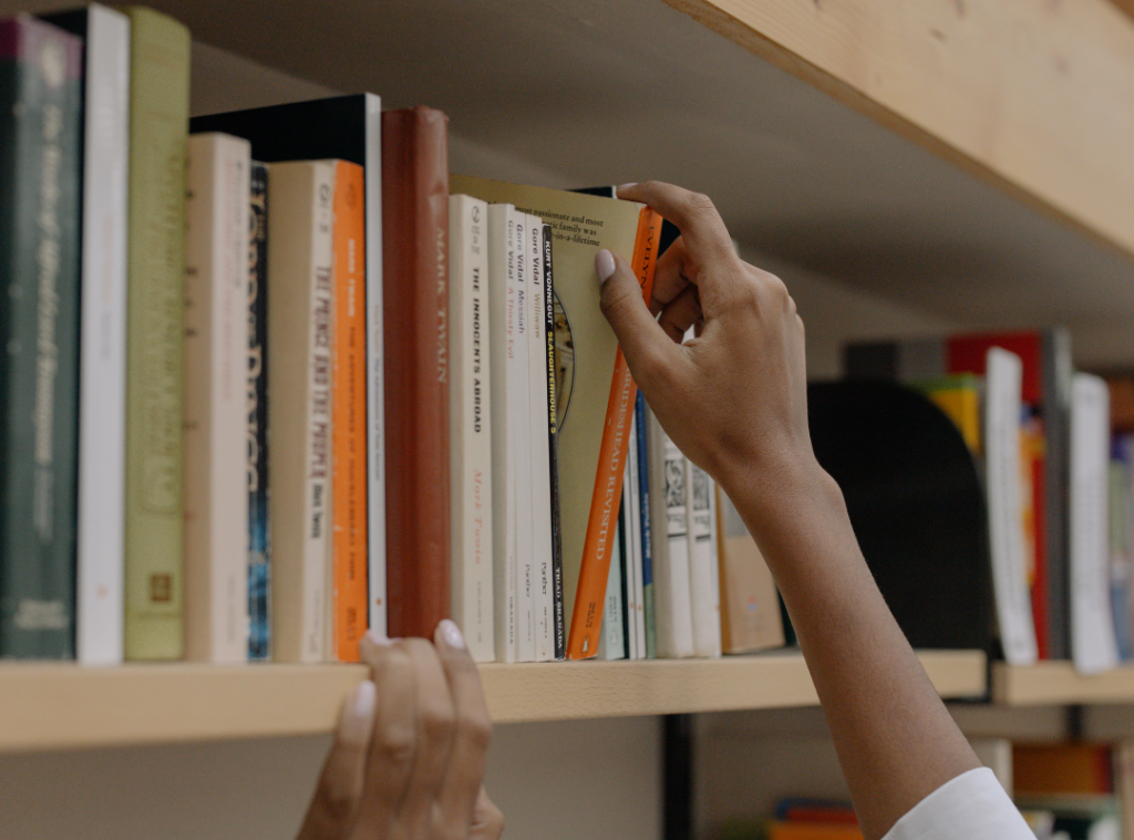 A woman's hand reaching up to retrieve a book from the book shelf in a library 