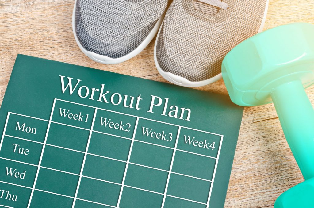 Workout plan on green board, with sneakers and dumbbells on wooden background.