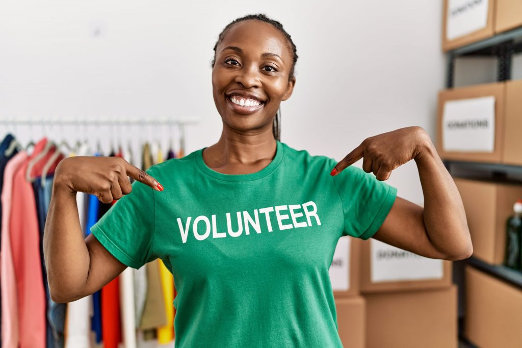 Young african american woman smiling happy pointing with finger to volunteer t shirt at charity center.